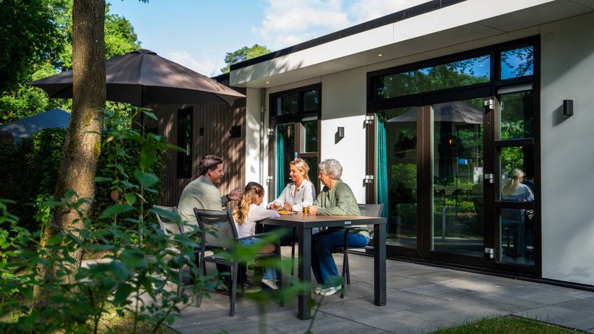 Vakantiegasten zitten ontspannen op het terras van hun vakantiewoning in recreatiepark 't Gelloo, omringd door groen.